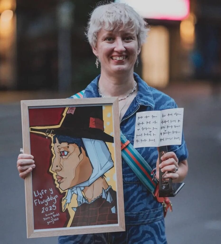 A person with short grey hair smiles whilst holding a framed artwork of a person in a hat and a sheet of stickers. The background is blurred.
